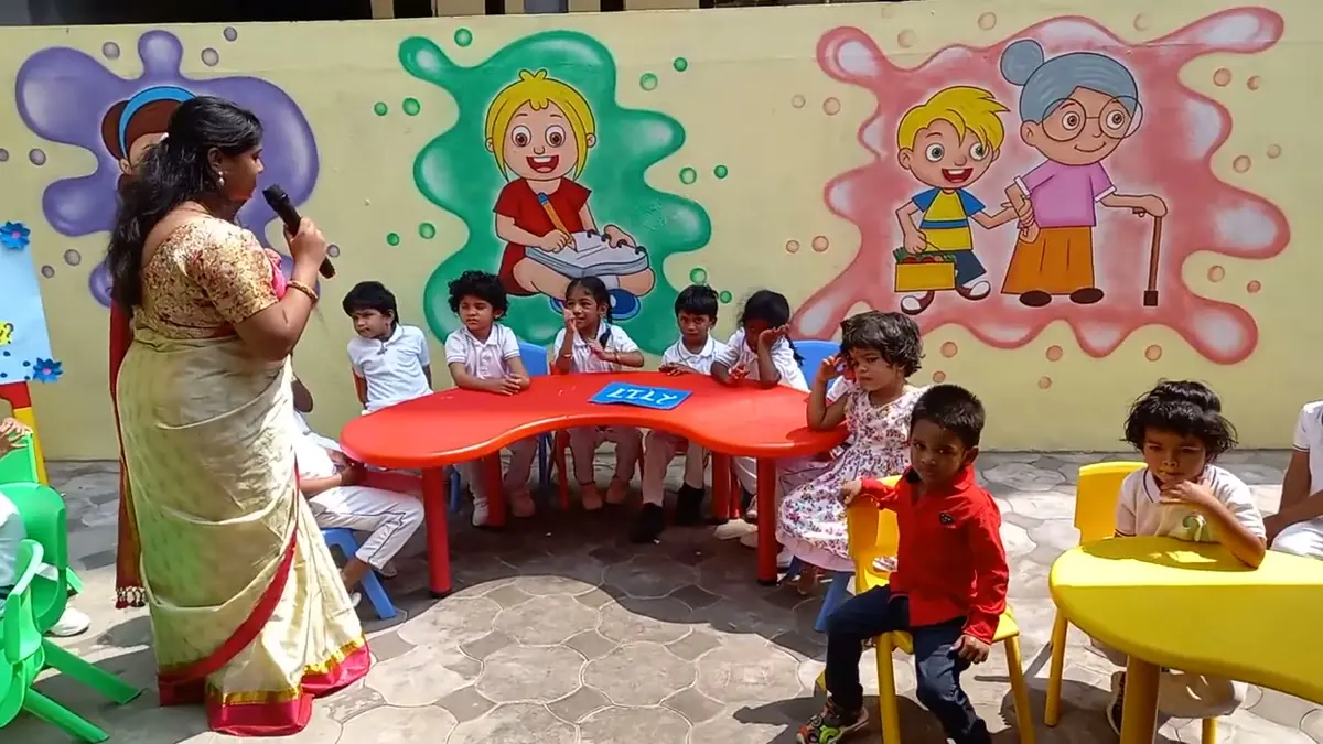 A teacher with a microphone sits with a group of children around a red table in the school's outdoor classroom, painted wall behind them.