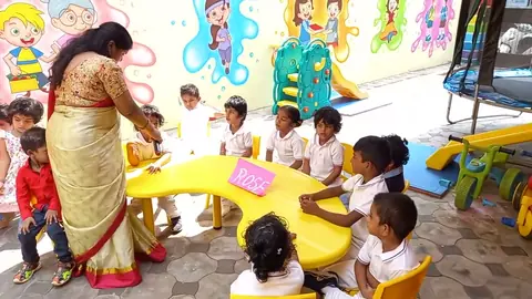 Teacher with a flashcard, children at a table, playground behind